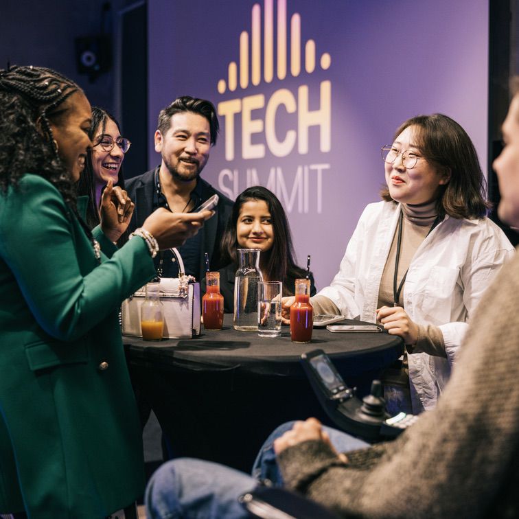 Diverse group of professionals networking at a tech summit event, engaged in conversation around a table with drinks